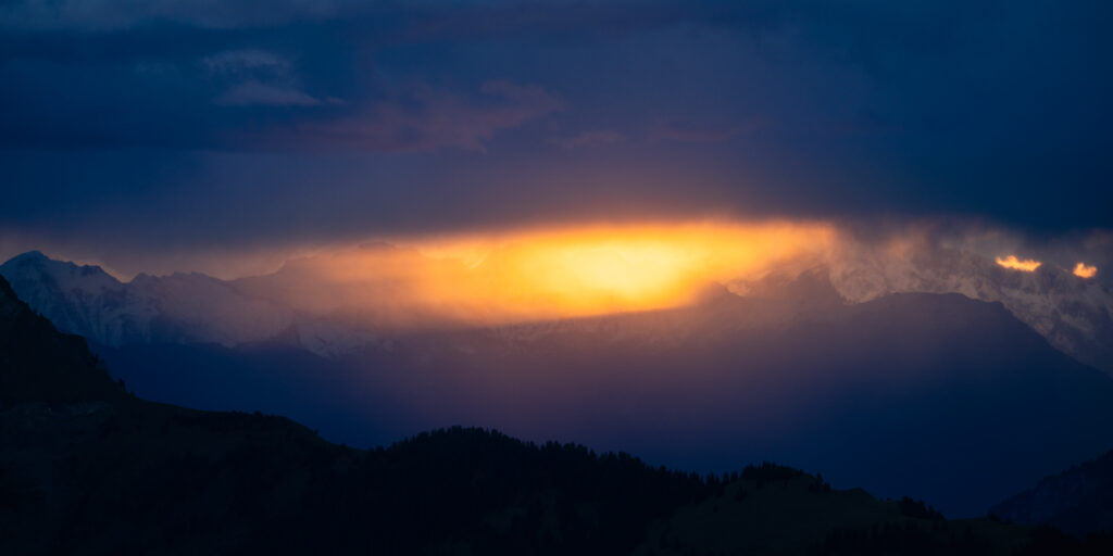 Düsterer Himmel mit Bergkulisse, bei dem die Sonne durch die Wolken drückt.